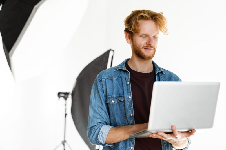 Academy of Acting Pleased handsome ginger guy smiling while working with laptop in studio indoors