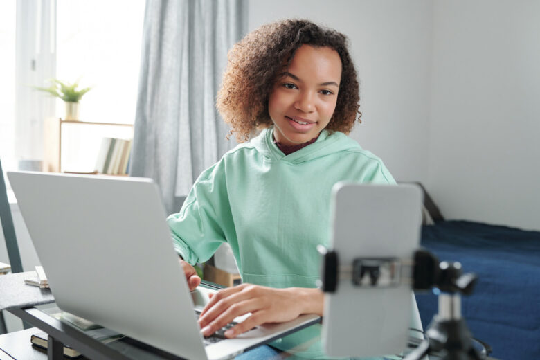 Academy of Acting Successful teenage student in casualwear sitting by table in bedroom and looking in smartphone camera while typing and recording video