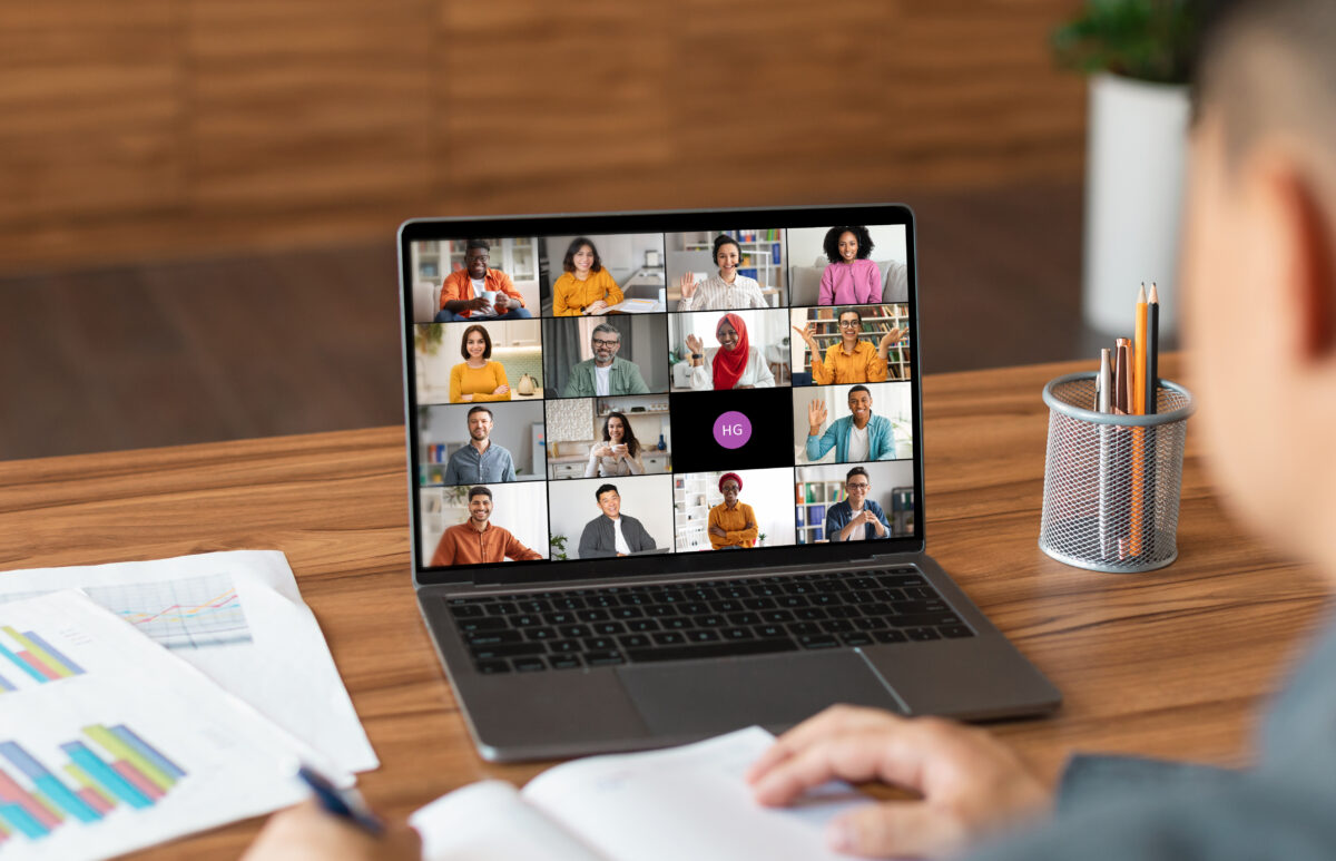 Academy of Acting A man sits at a desk in a home office and uses a laptop to participate in a video conference meeting. The laptop screen displays a grid of video windows featuring a diverse group of people